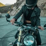 A biker in black leather gear poses confidently on a motorcycle on a winding mountain road.