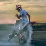 Motorcyclist performing a drift on pavement, creating smoke under a sunset sky.
