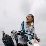 Woman posing on dirt bike with protective gear on a cloudy day.